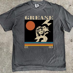 Può includere: T-shirt grigio scuro con grafica del film "Grease". Il design include la parola "GREASE", una coppia, un sole e l'anno "1978". La maglietta ha un aspetto vintage e usurato.