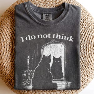 May include: A charcoal gray t-shirt featuring a white graphic of a black cat sitting in front of an ornate mirror. The text "I do not think" is printed above the cat. The shirt has a crew neckline and a Comfort Colors brand label at the neck.
