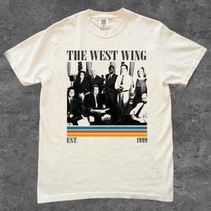 Puede incluir: Camiseta color crema con una imagen en blanco y negro de un grupo de personas. El texto "THE WEST WING" est&aacute; encima de la imagen, con "EST. 1999" debajo. La camiseta tiene cuello redondo y mangas cortas.