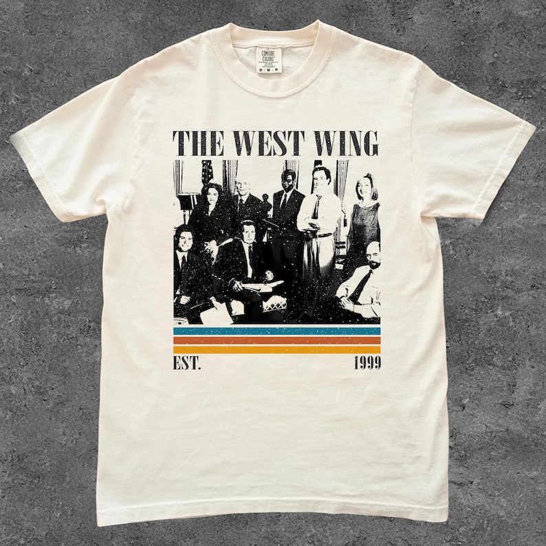 Puede incluir: Camiseta color crema con una imagen en blanco y negro de un grupo de personas. El texto "THE WEST WING" est&aacute; encima de la imagen, con "EST. 1999" debajo. La camiseta tiene cuello redondo y mangas cortas.