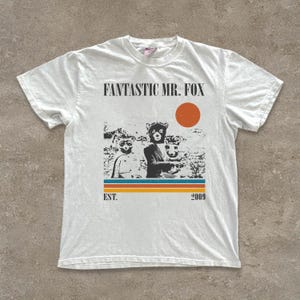 Può includere: T-shirt bianco con la scritta "FANTASTIC MR. FOX" sopra una grafica di tre personaggi animali e un cerchio rosso. Sotto la grafica ci sono strisce orizzontali e la scritta "EST. 2009".