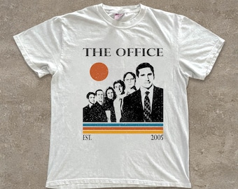 Może przedstawiać: Biała koszulka z grafiką z serialu "The Office". Projekt zawiera tytuł serialu, stylizowane słońce i linię postaci. Tekst "EST. 2005" jest również dołączony.