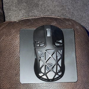 May include: A black computer mouse with a geometric design sits on a black mousepad. The mouse has a hollow, web-like pattern on its top surface. The mouse and pad are on a brown leather surface.