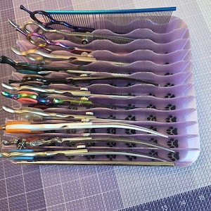 May include: A light purple plastic organiser tray with wavy slots holding multiple grooming scissors and a blue metal comb. The tray features a black paw print pattern at the base of each slot. A ruler is visible at the bottom, measuring up to 38 cm.
