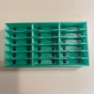 Puede incluir: Una bandeja de almacenamiento rectangular de color verde azulado con una cuadrícula de dieciocho compartimentos individuales. Cada compartimento tiene un borde frontal curvo y un pequeño icono de huella negra impreso en el fondo.
