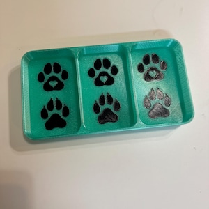 Puede incluir: Una bandeja rectangular turquesa con tres compartimentos, cada uno con huellas de patas negras. Algunas huellas tienen detalles en forma de corazón. La bandeja está diseñada para dueños de mascotas.