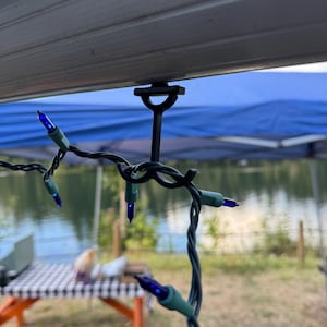 May include: A black plastic T-shaped hook clip attached to a metal frame, supporting a string of dark green wire with small blue LED light bulbs. The hook features a curved design to hold the light strand securely in place along the structure.