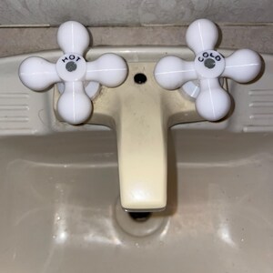 May include: Close-up of a bathroom sink with a cream-colored basin and faucet. The white faucet handles are in a cross shape, labeled "HOT" and "COLD". The sink is clean and appears to be in good condition.