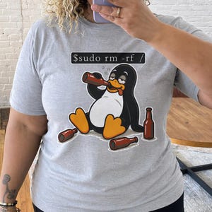 Pode incluir: Camiseta cinza claro com um desenho de um pinguim bebendo de uma garrafa marrom. O pinguim é preto e branco com pés laranja e um bico amarelo. O texto "$sudo rm -rf /" está acima do pinguim.