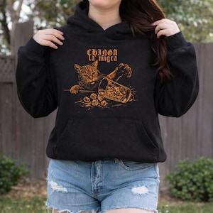 Puede incluir: Sudadera con capucha negra con un gr&aacute;fico de un gato alcanzando un vaso de whisky y cubitos de hielo. El texto "Chinga la migra" est&aacute; encima del gato. La sudadera tiene un bolsillo delantero y una capucha con cord&oacute;n.