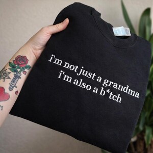 以下が含まれることがあります： 白い文字で「I'm not just a grandma, I'm also a b*tch」と書かれた黒いスウェットシャツ。 バラと注射器のタトゥーが入った手でスウェットシャツが持たれています。