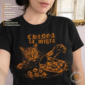 May include: Black t-shirt featuring an orange illustration of a cat, a tipped-over glass of ice cubes, and the text "CHINGA la migra." The artwork is hand-drawn and copyrighted. The shirt supports independent art and original creators.