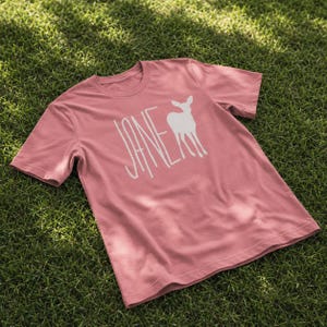 Peut inclure: Un t-shirt à manches courtes rose poudré avec une impression graphique blanche sur le devant. Le motif se compose du mot JANE écrit dans une police haute et fine, accompagné d'une silhouette blanche de cerf ou de faon debout de face.