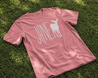 Può includere: Una t-shirt a maniche corte color rosa polvere con una stampa grafica bianca sul davanti. Il design presenta la parola JANE scritta con un carattere alto e sottile accanto alla sagoma bianca di un cervo o cerbiatto in piedi di fronte.