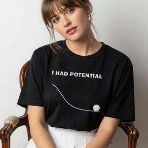 Könnte beinhalten: Eine weiße Keramik-Tasse mit schwarzem Henkel und dem Text "But First, Coffee" in schwarzer, fetter Schrift. Die Tasse steht auf einer Holzoberfläche.