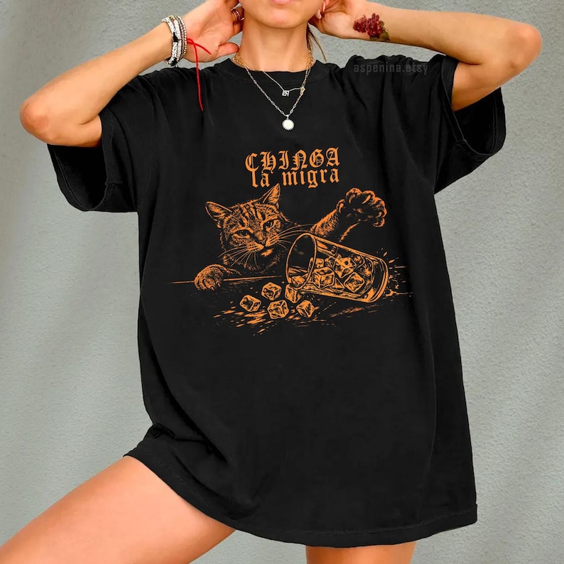 Puede incluir: Camiseta negra con un gr&aacute;fico naranja de un gato volcando un vaso de cubitos de hielo. El texto "Chinga la migra" est&aacute; encima del gato. La camiseta tiene mangas cortas y un corte holgado.