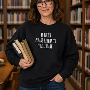 May include: Black sweatshirt with the text "IF FOUND PLEASE RETURN TO THE LIBRARY" in white. The sweatshirt is worn by a person holding several books. The background shows bookshelves filled with books.