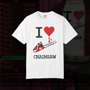 May include: White t-shirt featuring a graphic design. The design includes the text "I", a red heart with blood dripping, a red chainsaw, and the word "CHAINSAW". The shirt is a tribute to the 1983 American splatter movie.
