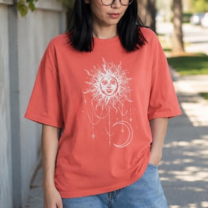 Puede incluir: Una camiseta de manga corta color coral con un estampado gráfico blanco de un sol con rostro humano, rodeado de rayos, cadenas colgantes y una luna creciente, centrado en la parte delantera de la prenda.