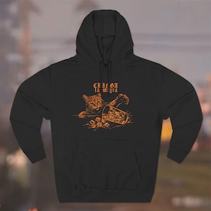 Puede incluir: Sudadera con capucha negra con una ilustraci&oacute;n naranja de un gato y un vaso con cubitos de hielo. El texto "CLICA la migra" est&aacute; encima del gato. La sudadera tiene una capucha con cord&oacute;n y un bolsillo delantero.