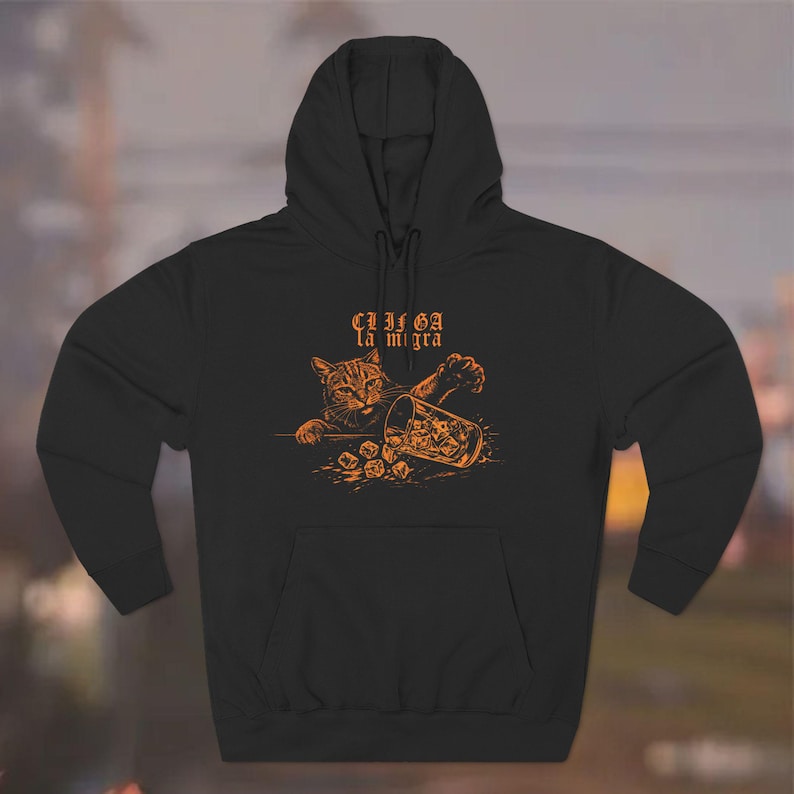 Puede incluir: Sudadera con capucha negra con una ilustraci&oacute;n naranja de un gato y un vaso con cubitos de hielo. El texto "CLICA la migra" est&aacute; encima del gato. La sudadera tiene una capucha con cord&oacute;n y un bolsillo delantero.