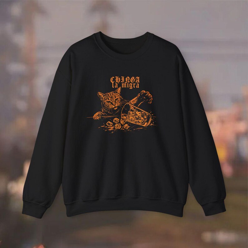 Puede incluir: Sudadera negra con un gr&aacute;fico naranja de un gato que alcanza un vaso con dados. El texto "Chinga la migra" est&aacute; encima del gato. El gato es marr&oacute;n y naranja. La sudadera es de cuello redondo.