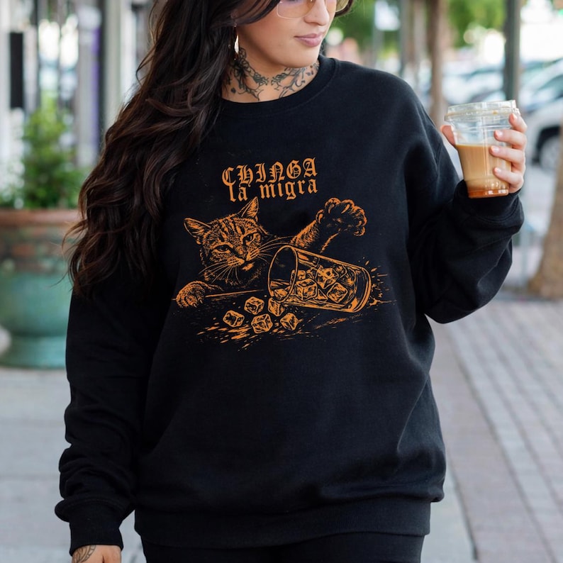 Puede incluir: Sudadera negra con un gr&aacute;fico naranja de un gato volcando un vaso de hielo. El texto "Chinga la migra" est&aacute; encima del gato. El gato y el vaso est&aacute;n detallados con sombreado naranja.