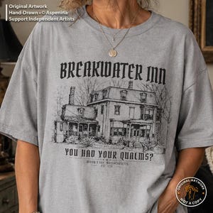 May include: A heather gray t-shirt featuring a black ink-style illustration of a large house. The text reads BREAKWATER INN, YOU HAD YOUR QUALMS?, WIDOW'S BAY, MASSACHUSETTS, EST. 1731. The design includes bare trees and architectural details.
