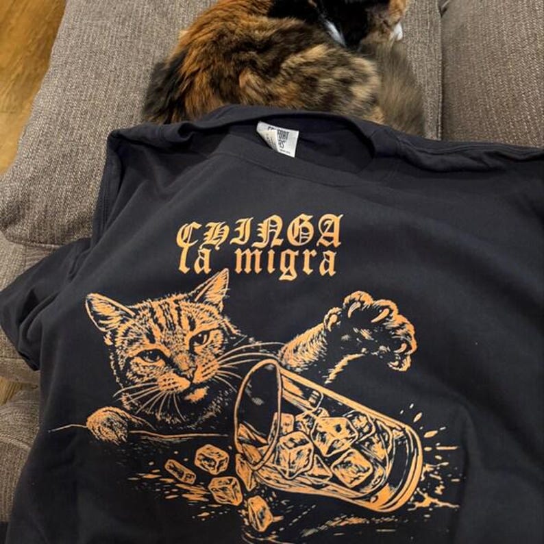 Puede incluir: Camiseta negra con un gr&aacute;fico naranja de un gato volcando un vaso de hielo. El texto "CHINGA la migra" est&aacute; encima de la ilustraci&oacute;n del gato. Un gato calic&oacute; descansa en el sof&aacute; detr&aacute;s de la camiseta.