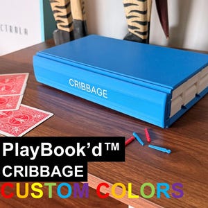 May include: A bright blue rectangular cribbage board shaped like a book with the word CRIBGAGE printed in white on the spine. The set includes red and blue plastic pegs and a deck of red playing cards. The text PlayBook'd CRIBAGE CUSTOM COLORS is visible.