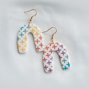 May include: A pair of white, arch-shaped earrings with gold-colored hooks. Each earring is decorated with colorful plus signs in a rainbow of hues, including yellow, orange, red, purple, blue, and green. The earrings are displayed on a white surface.