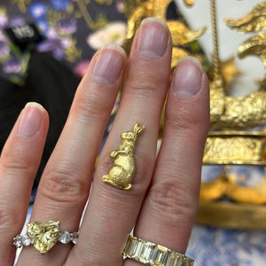 May include: A gold-colored bunny ring is displayed on a finger, alongside two other rings. One ring features a large yellow heart-shaped stone, and the other has a band of rectangular stones. The bunny ring is the focal point.