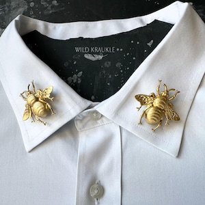 Peut inclure: Deux épingles de col en forme d'abeille dorée sur une chemise blanche. Les épingles présentent des ailes et des segments de corps détaillés. La chemise a un col boutonné et une patte de boutonnage. Le fond est sombre avec le texte "WILD KRAUKLE".