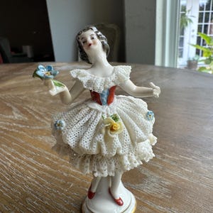 May include: A porcelain figurine of a person in a ruffled white dress with a textured lace-like pattern, featuring a red and blue bodice, red shoes, and a gold-trimmed base. The figure holds a small blue flower and has a yellow rose on the skirt.