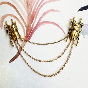 Puede incluir: Broche de metal dorado con dos figuras de escarabajos unidas por tres delicadas cadenas. Los escarabajos tienen cuerpos texturizados y detallados sobre un fondo de acuarela con tonos rosas y azules. Un accesorio único.