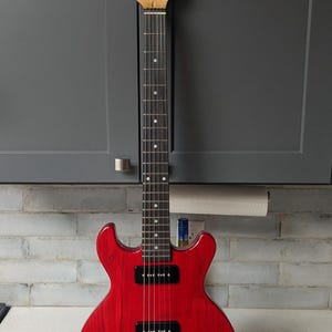 Pode incluir: Uma guitarra elétrica vermelha com braço e headstock de madeira. A guitarra tem captadores pretos, botões e uma escala com incrustações de pontos brancos. O headstock tem o nome da marca "Duval".
