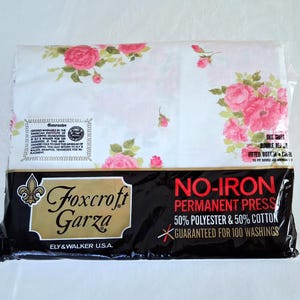 May include: A packaged set of Foxcroft Garza bed sheets. The sheets are white with a floral pattern of pink roses and green leaves. The packaging is black with gold lettering and the words "NO-IRON PERMANENT PRESS".