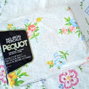 May include: A packaged double flat sheet with a floral pattern, featuring pink, blue, and yellow flowers on a white background. The packaging reads "PEQUOT" and "NO-IRON PERCALE". The sheet is designed for a 54x76 inch mattress.