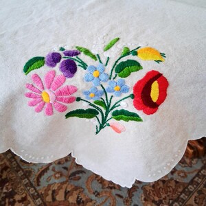 May include: White tablecloth with a scalloped edge, embroidered with a colorful floral bouquet. The bouquet features pink, purple, blue, red, and yellow flowers with green leaves. The embroidery is detailed and vibrant.