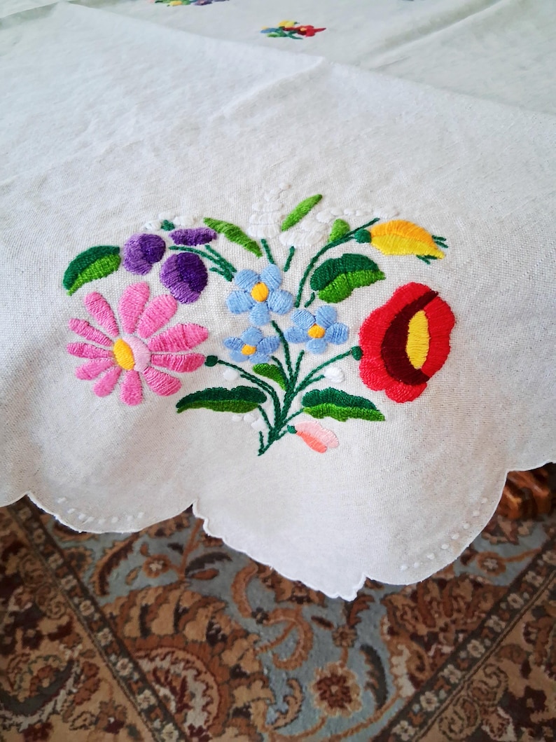 May include: White tablecloth with a scalloped edge, embroidered with a colorful floral bouquet. The bouquet features pink, purple, blue, red, and yellow flowers with green leaves. The embroidery is detailed and vibrant.