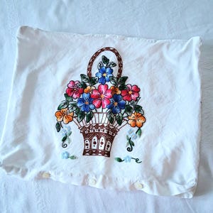 May include: A white rectangular pillowcase featuring a central embroidered design of a brown woven basket filled with vibrant red, blue, orange, and yellow flowers with green leaves. The bottom edge includes three small white buttons for closure.