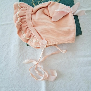 May include: A vintage baby bonnet in a soft peach corduroy. The bonnet features a ruffled edge with delicate white lace trim and a satin ribbon bow. The bonnet has long satin ties for securing under the chin. A teal book with gold lettering is in the background.