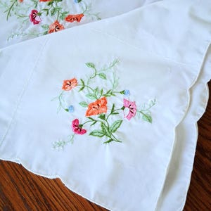 May include: White embroidered pillowcases with colorful floral designs. The embroidery features orange, pink, and purple flowers with green leaves. The pillowcases have a scalloped edge and are made of a soft, white fabric.