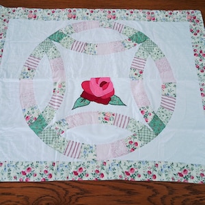 May include: A handmade quilt featuring a central rose applique. The quilt has a white background with a double wedding ring design in pink and green patchwork. The border is floral with pink roses.