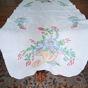 May include: White embroidered table runner featuring a floral design. The embroidery includes blue flowers, green leaves, and red berries, arranged around a cornucopia. The runner is placed on a wooden table.