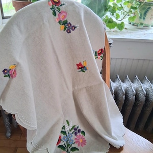 May include: A cream-colored, embroidered tablecloth with a scalloped edge. The tablecloth features colorful floral embroidery in pink, red, blue, and purple. The cloth is draped over a wooden chair.