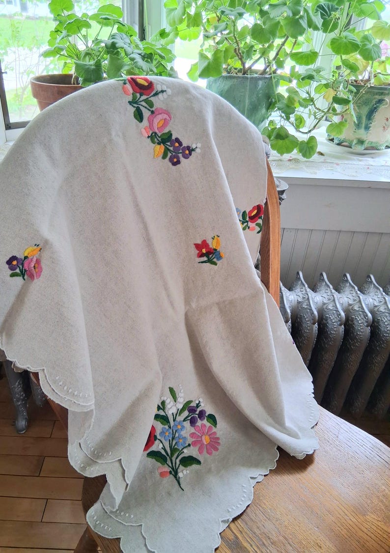 May include: A cream-colored, embroidered tablecloth with a scalloped edge. The tablecloth features colorful floral embroidery in pink, red, blue, and purple. The cloth is draped over a wooden chair.