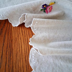 May include: Off-white linen fabric with a scalloped edge and embroidered floral details. The embroidery features pink, purple, yellow, and green thread. The fabric is draped over a wooden surface.