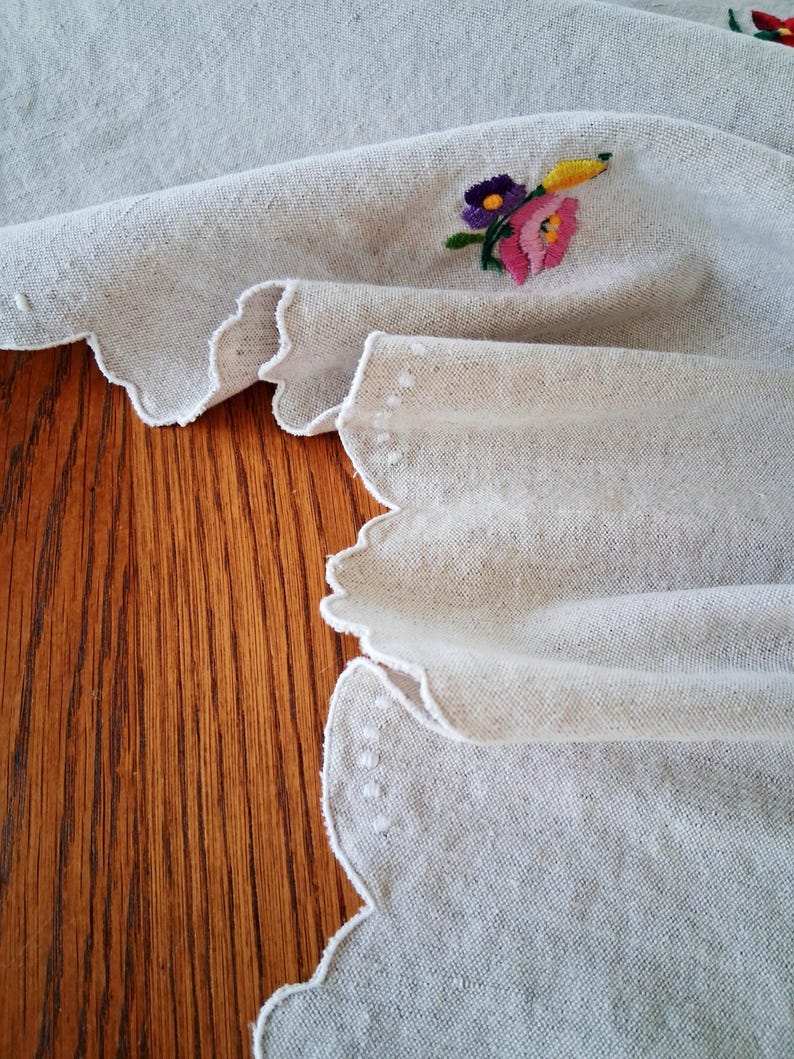 May include: Off-white linen fabric with a scalloped edge and embroidered floral details. The embroidery features pink, purple, yellow, and green thread. The fabric is draped over a wooden surface.