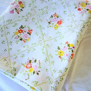 May include: A white fabric featuring a repeating pattern of small floral bouquets in shades of pink, yellow, and orange. The bouquets are connected by a light green lattice design of thin, leafy vines that create a diamond-shaped grid across the surface.